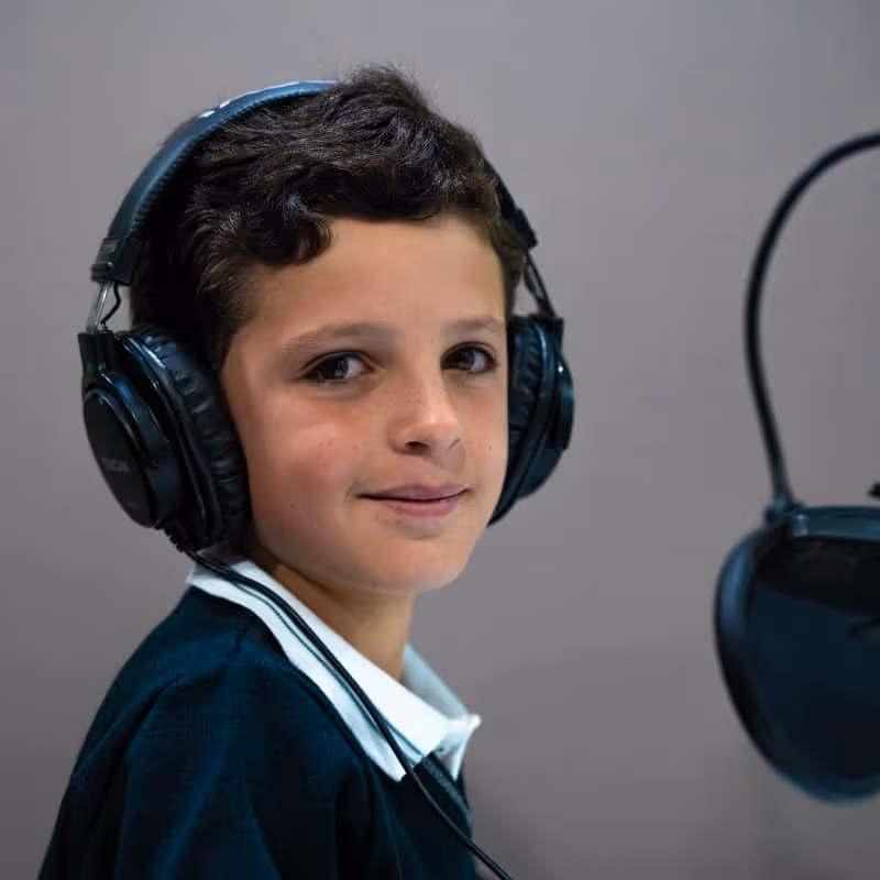 Un joven con grandes auriculares se sienta frente a un micrófono y sonríe ligeramente a la cámara en un estudio de grabación.